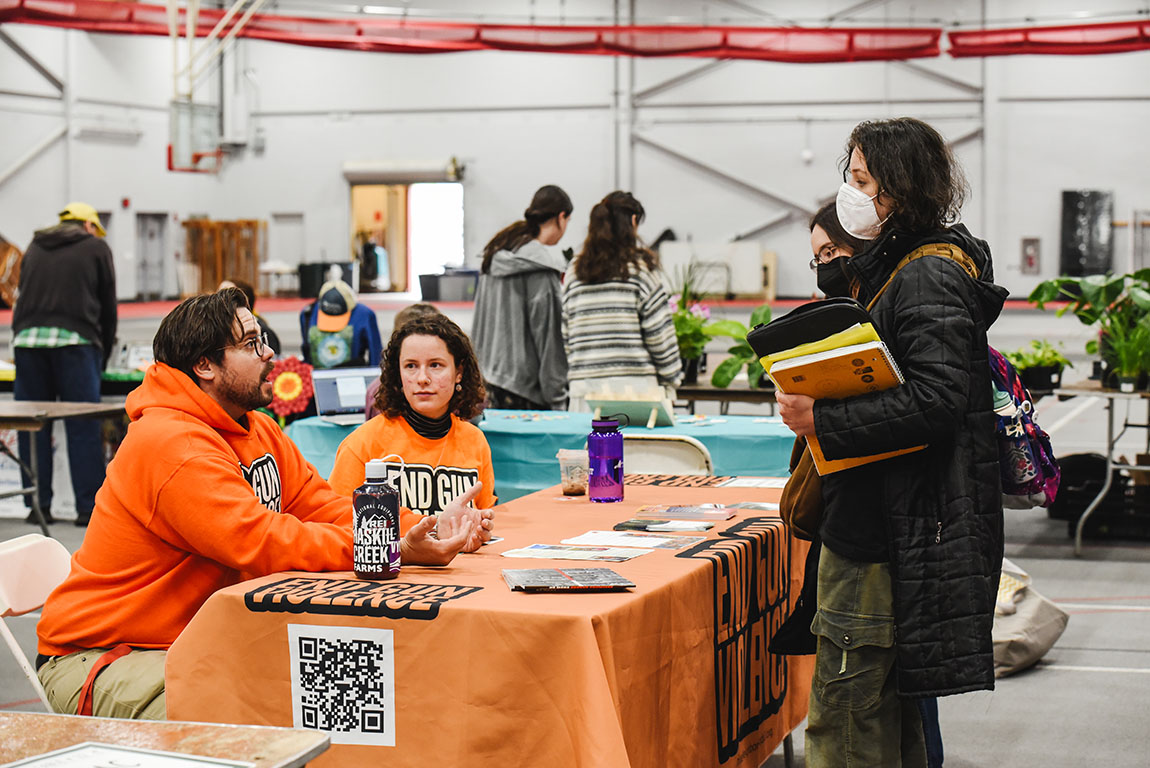 Two young adults in orange T-shirts that say END GUN VIOLENCE sit at a table and speak with two young people who are standing